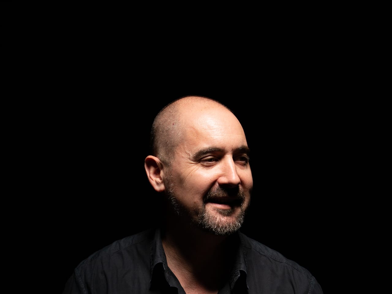 Huw Davies, smiling and looking off camera, lit against a black background.